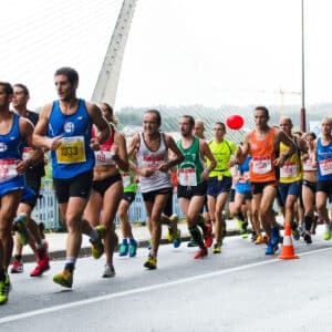 A group of runners in numbered bibs participate in a 13.1 Half Marathon race on a city street near a cable-stayed bridge, with spectators behind barriers.