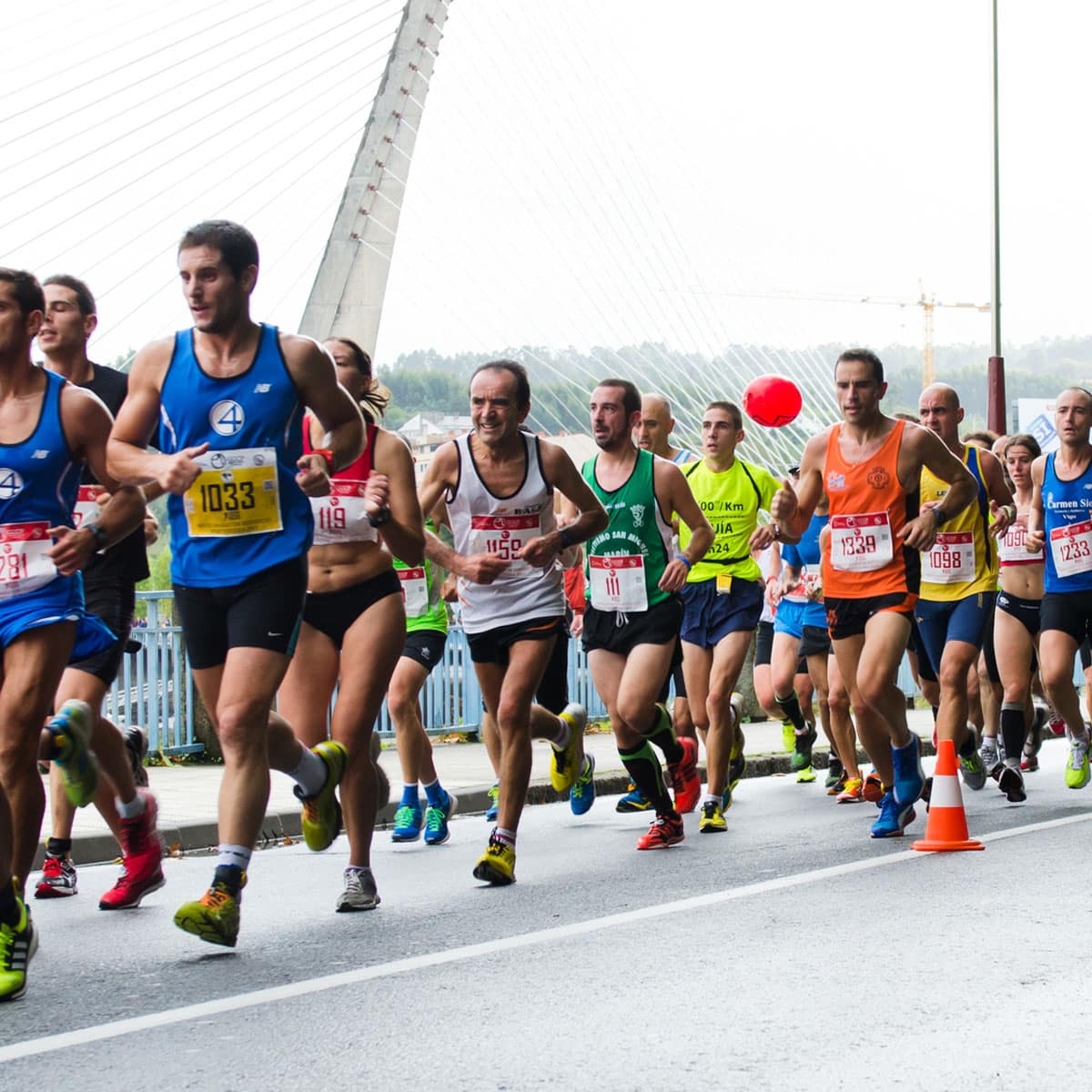 A group of runners in numbered bibs participate in a 13.1 Half Marathon race on a city street near a cable-stayed bridge, with spectators behind barriers.