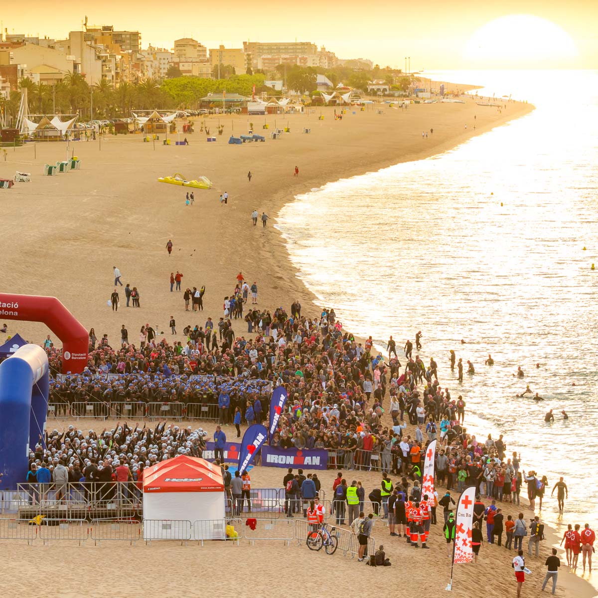 A large crowd gathers at sunrise on a sandy beach, with athletes ready to start an Ironman event by swimming 2.4 miles. City buildings rise in the background as the ocean reflects the sun.