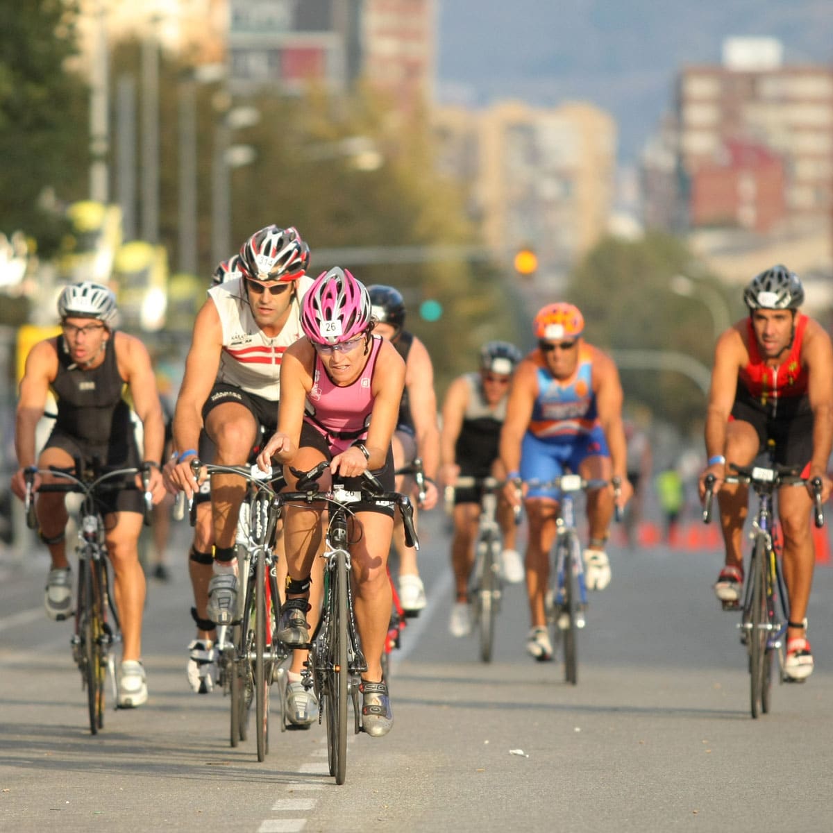 A group of cyclists wearing helmets and athletic gear ride closely together on a city street during a 70.3 triathlon race covering 56 miles. The leading cyclist is sporting a pink helmet.