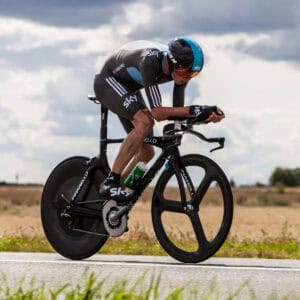 A cyclist in a black and blue Team Sky team kit rides a time trial bike with aero bars on a rural road conveying the concept of using aero bars to increase aerodynamic efficiency in a TT event.