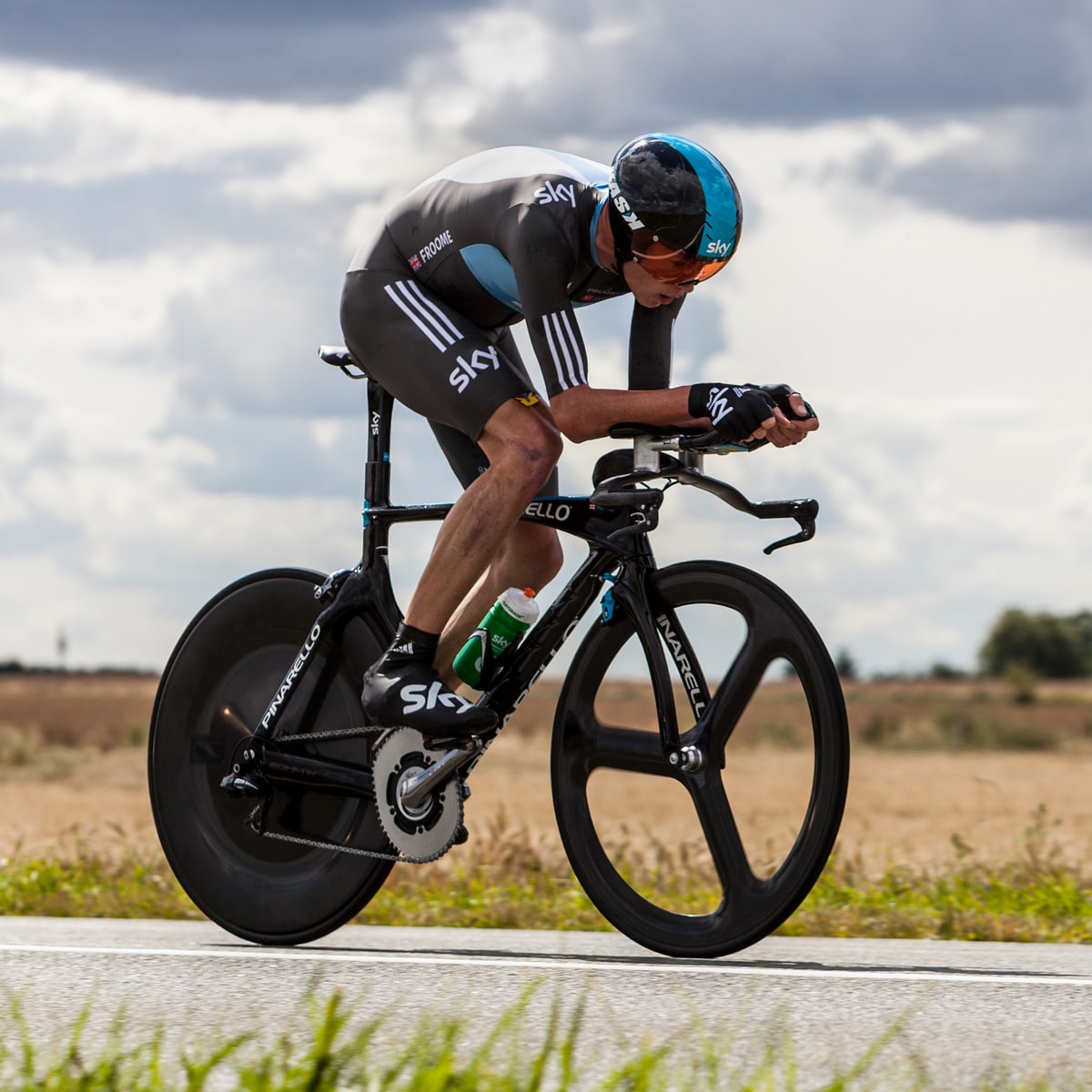 A cyclist in a black and blue Team Sky team kit rides a time trial bike with aero bars on a rural road conveying the concept of using aero bars to increase aerodynamic efficiency in a TT event.