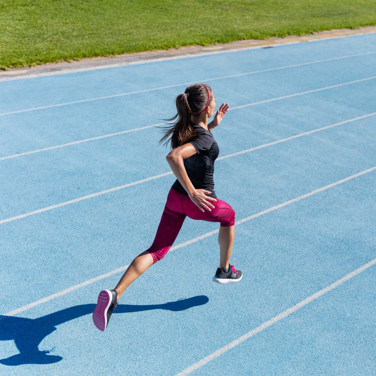 A woman wearing a black top and red leggings is sprinting on a blue running track, with grass visible in the background. She is running at full speed, mid-stride, with her hair in a ponytail, resembling the concept of anaerobic efforts.