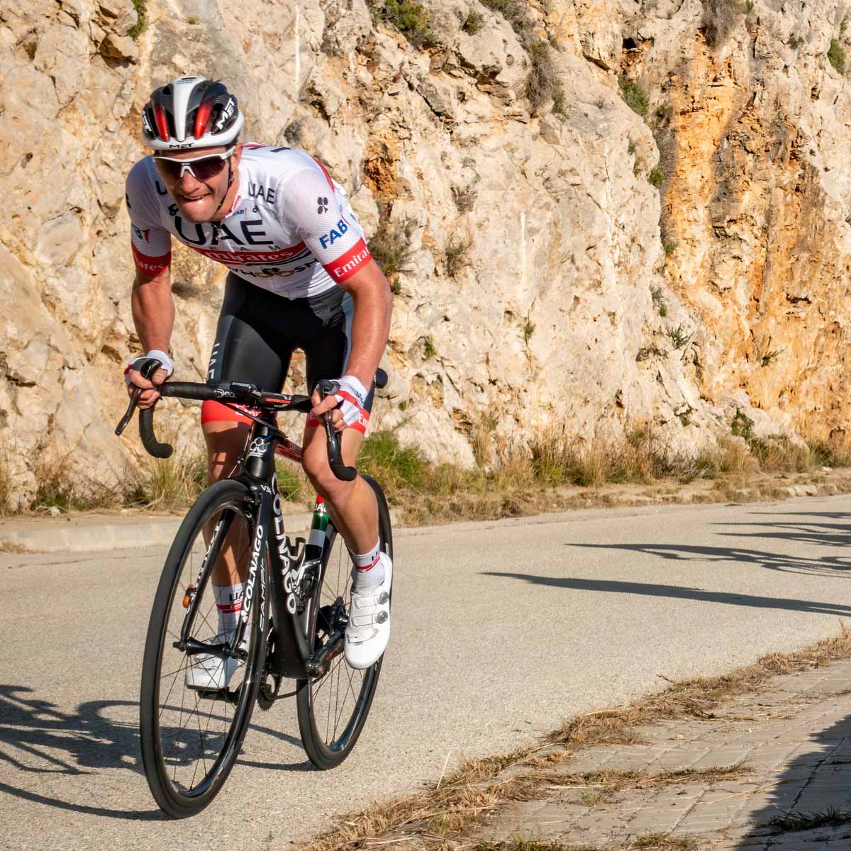 A cyclist in a white and red UAE team jersey and helmet rides uphill on a road next to a rocky cliff, appearing focused and determined under sunny weather.