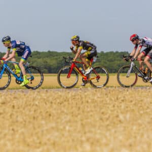 Three cyclists ride racing bikes on a rural road beside a wheat field, with trees and clear sky in the background. The cyclists wear colorful jerseys and helmets.