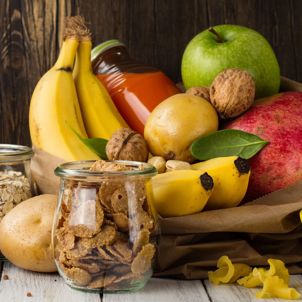 A variety of fresh fruits, nuts, cereal, and a jar of orange juice are arranged together on a wooden surface. The image conveys the concept of carbohydrates as high-octane fuel for endurance sports performance.