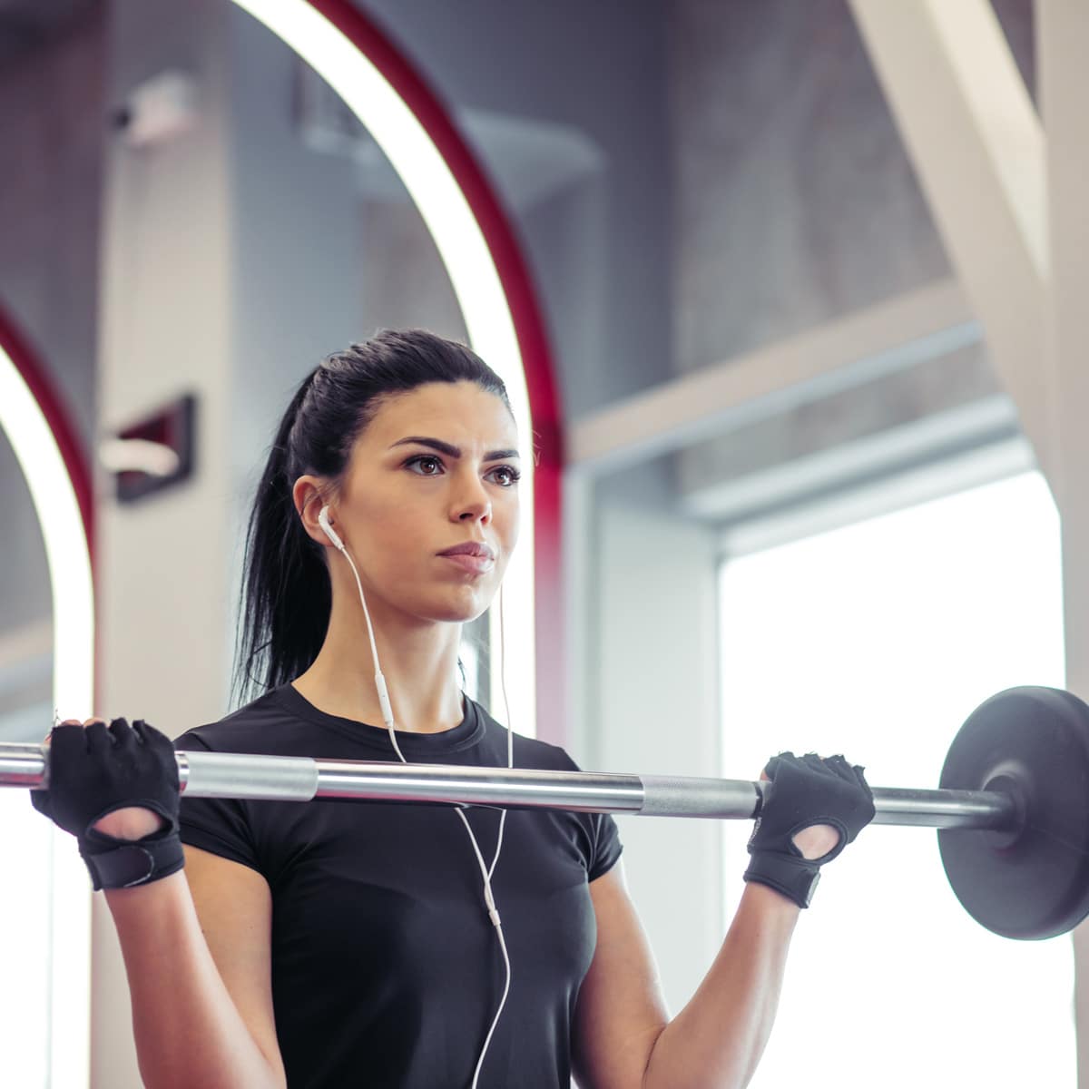 A woman wearing black workout gloves and earphones lifts a barbell in a gym, looking focused. She has long dark hair tied back and is dressed in a black athletic shirt. Bright gym lights and equipment are visible in the background.