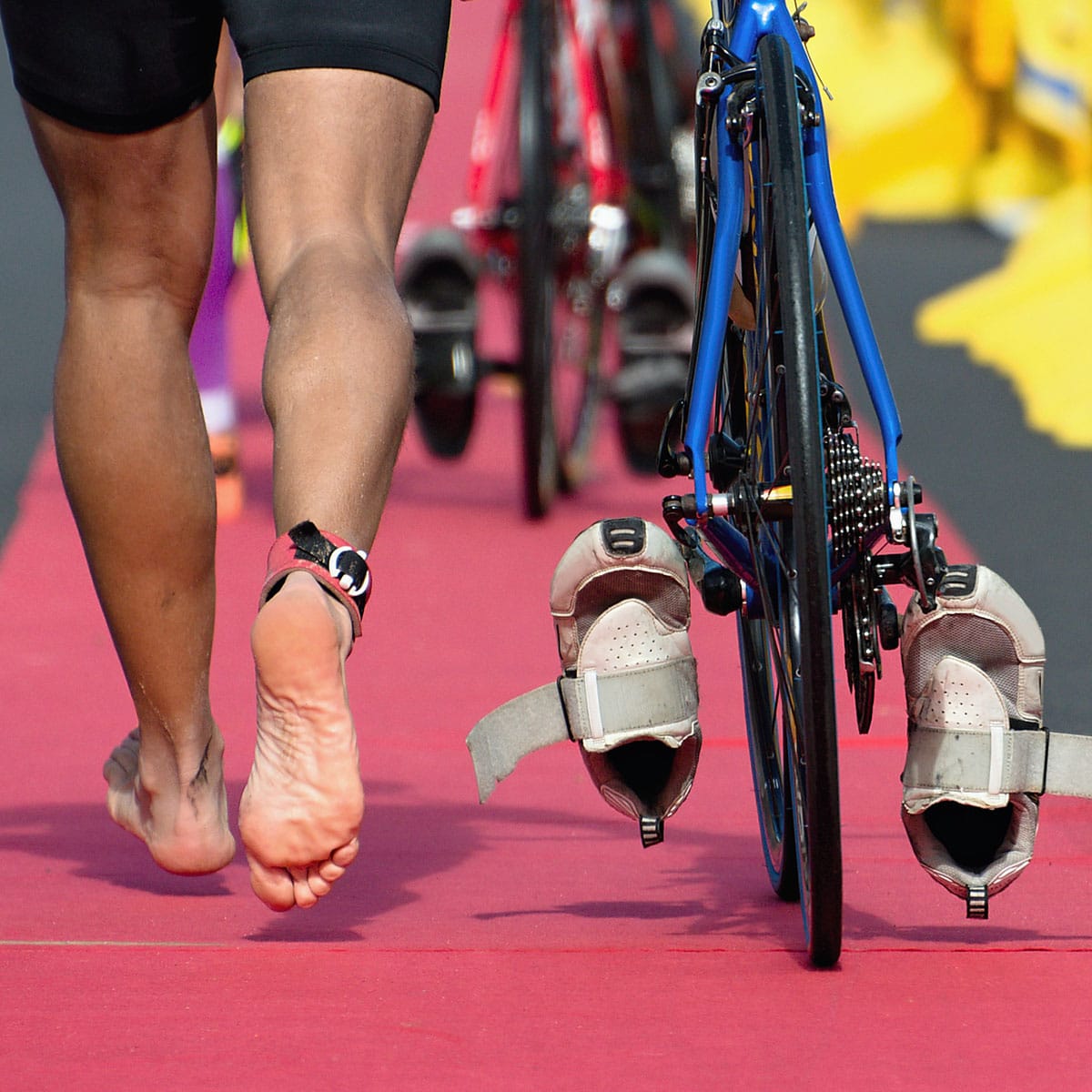 A barefoot athlete runs next to a blue bicycle with shoes clipped to the pedals on a red track during a triathlon transition.