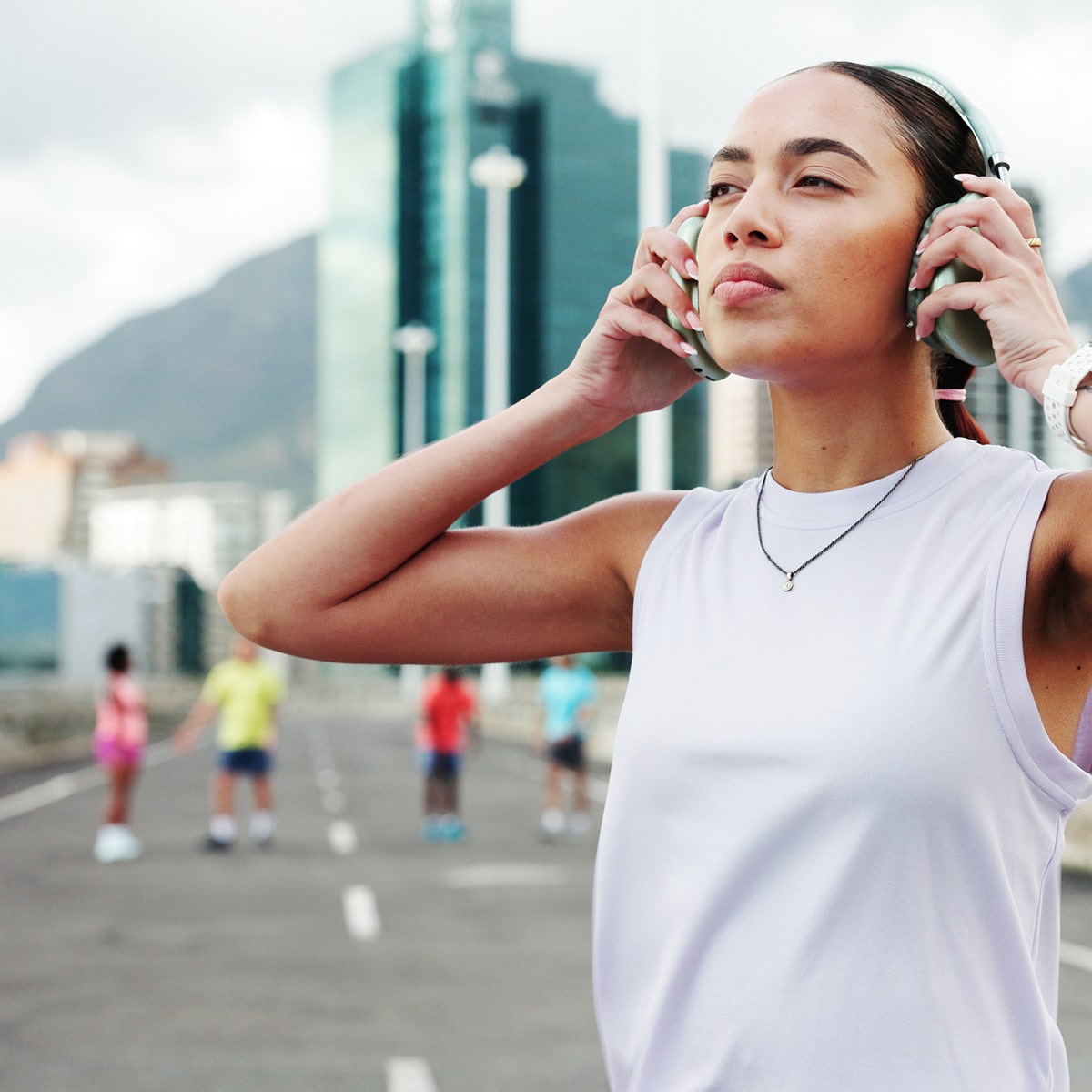 A woman stands outdoors wearing headphones, appearing focused or meditative. Blurry people exercise in the background on a city street near tall buildings and a mountain.