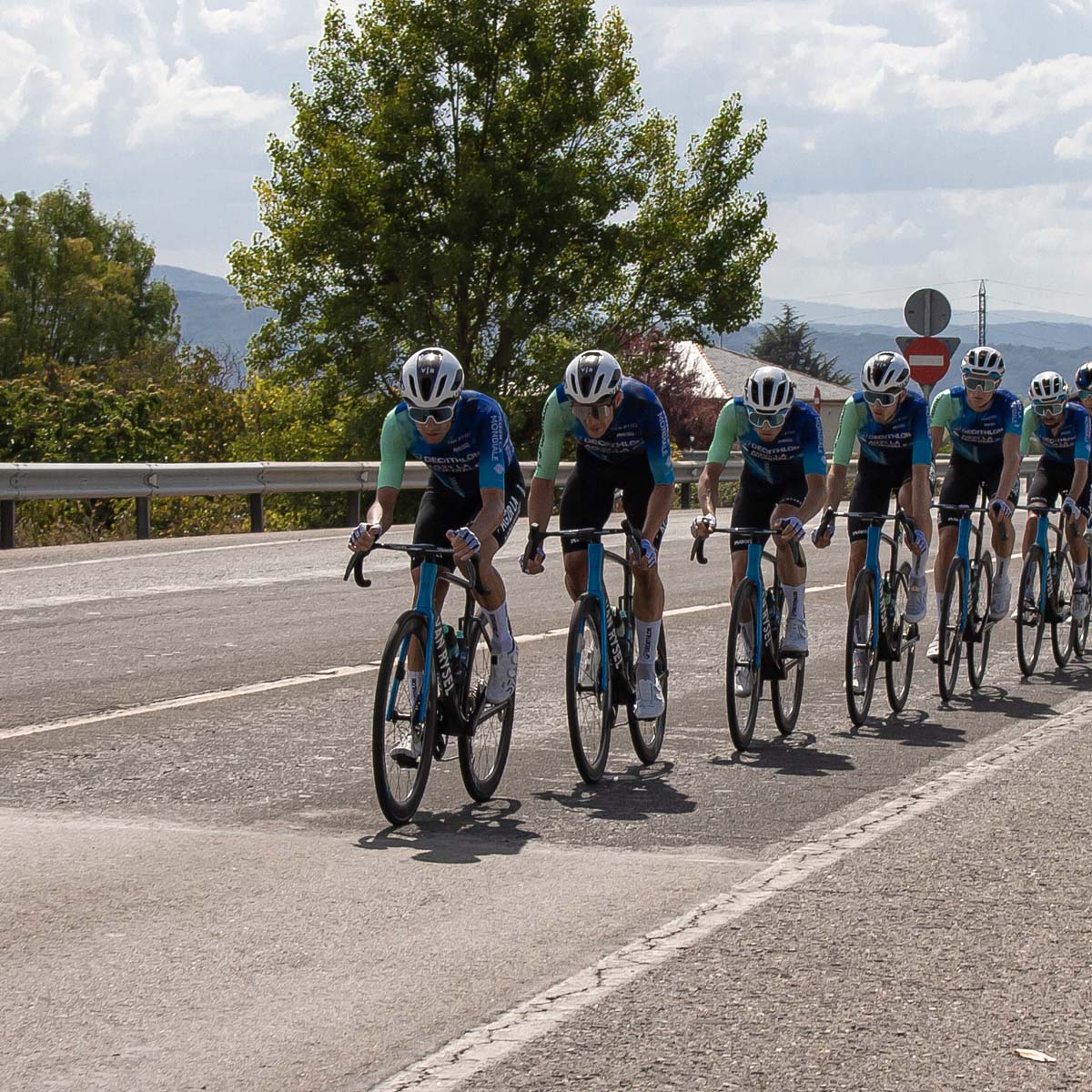A group of cyclists in matching uniforms ride in a staggered echelon formation on a sunny road, with trees and mountains behind them.