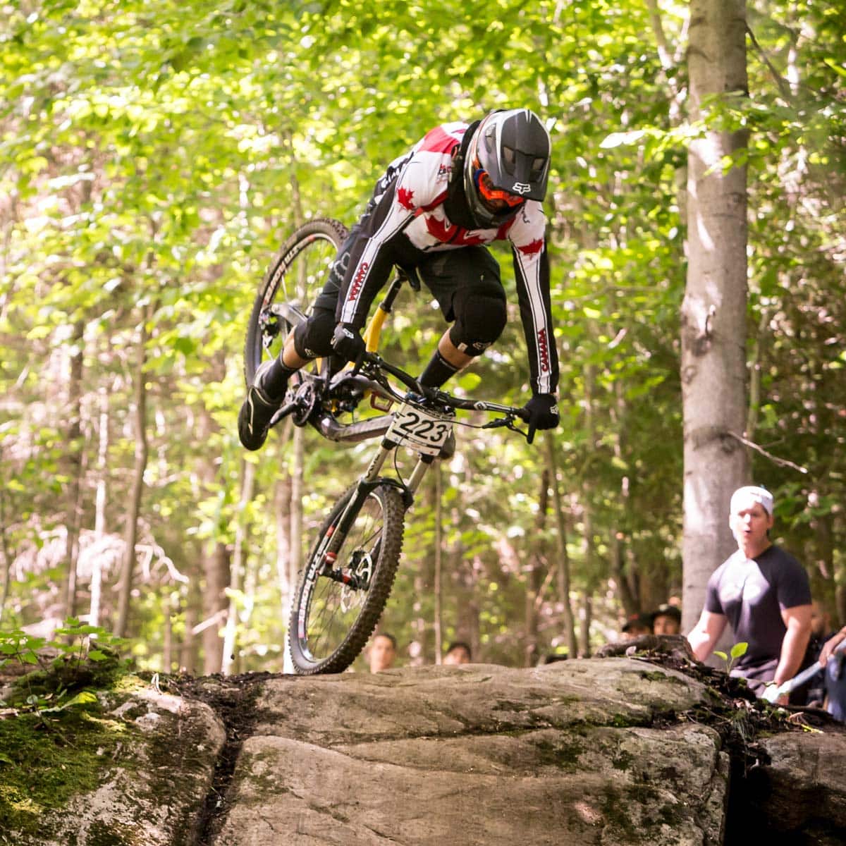 A mountain biker wearing a helmet and a red-and-white jersey flips over the handlebars on a forest trail during a race, with sunlight filtering through the trees. The bike’s front wheel is off the ground and the rider is airborne.