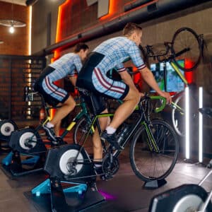 Two cyclists wearing matching jerseys ride stationary bikes indoors. The gym features modern equipment, wall-mounted bicycles, and an orange-lit, tech-filled atmosphere.