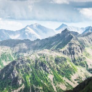 A scenic view of rugged, green and rocky mountain peaks under a partly cloudy sky, with distant, misty mountains in the background.