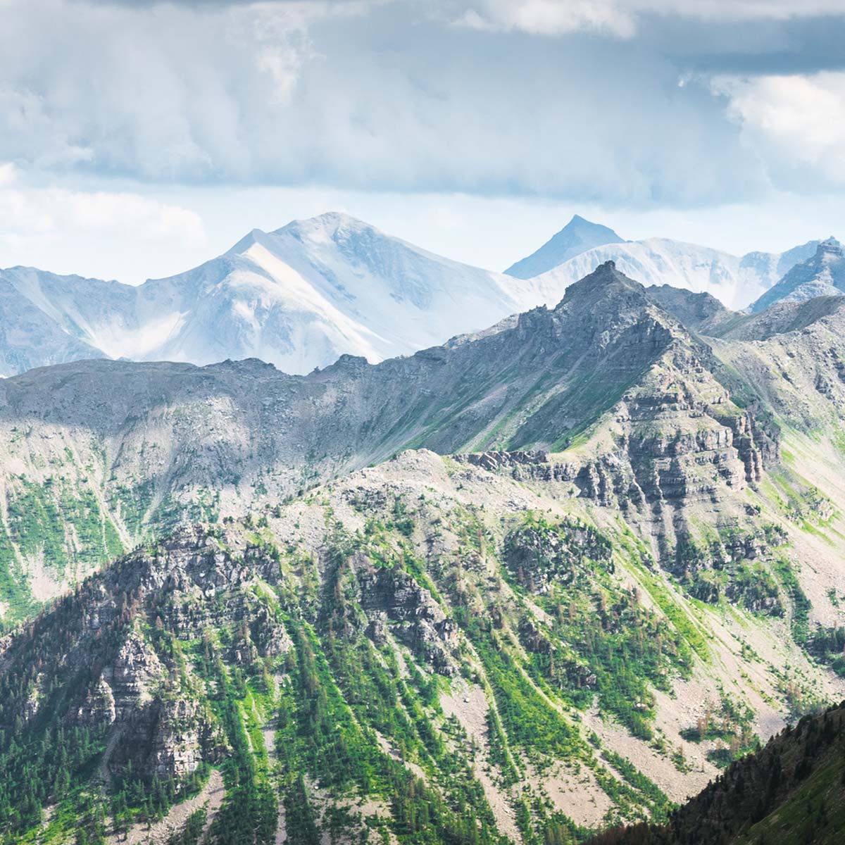 A scenic view of rugged, green and rocky mountain peaks under a partly cloudy sky, with distant, misty mountains in the background.