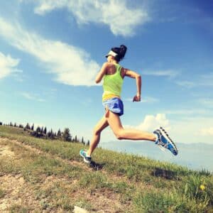 A woman in athletic wear and a visor runs outdoors during a Fartlek run along a grassy trail, with mountains, trees, and a blue sky in the background.