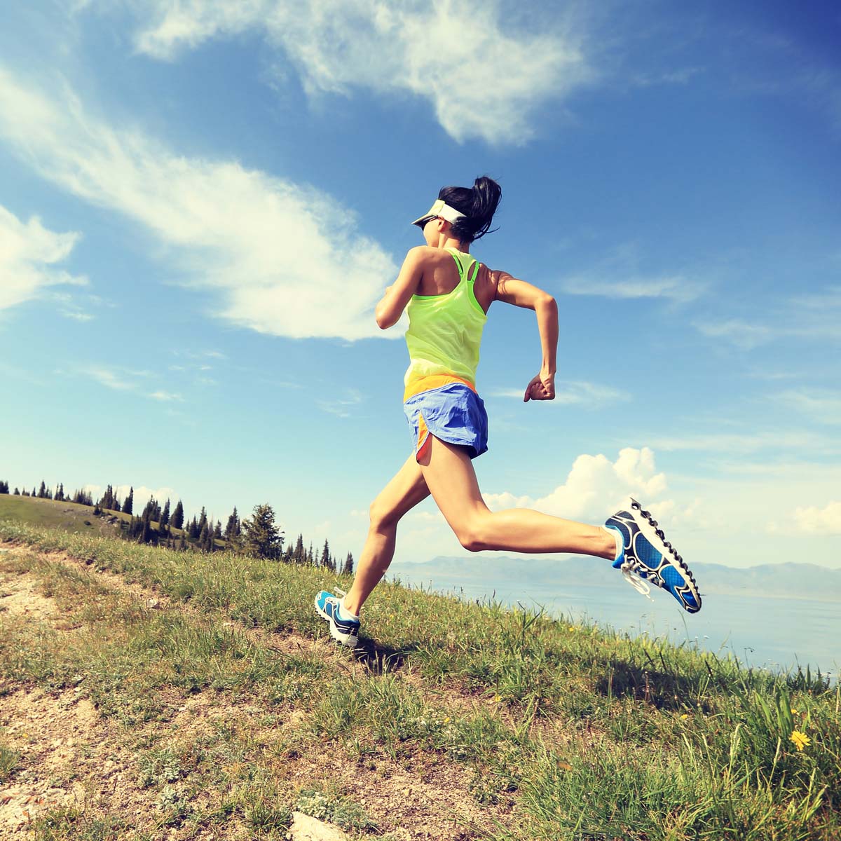 A woman in athletic wear and a visor runs outdoors during a Fartlek run along a grassy trail, with mountains, trees, and a blue sky in the background.
