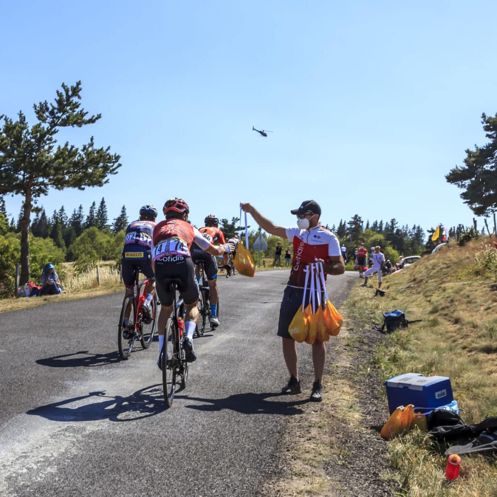 Cyclists ride on a tree-lined road as a team member hands out food bags in the feed zone. A helicopter flies in the blue sky above, while spectators watch from the grassy roadside.