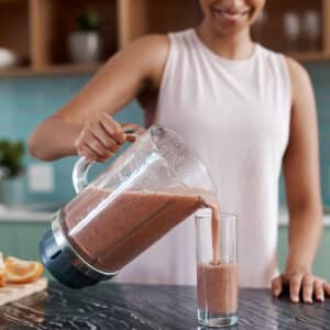 A woman in a sleeveless top pours a thick, brown smoothie from a blender pitcher into a glass, standing at a kitchen counter with fruit in the background—perfect for post workout nutrition and supporting muscle recovery.