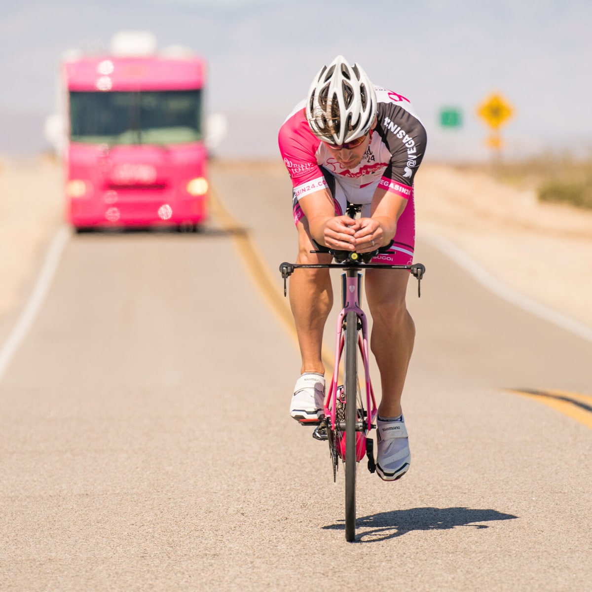 A cyclist in pink gear rides on an open road, followed by a pink support RV during a Vegas In 24 ultra-cycling challenge, covering one of the four routes required to achieve the Grand Slam in the Hall of Fame.