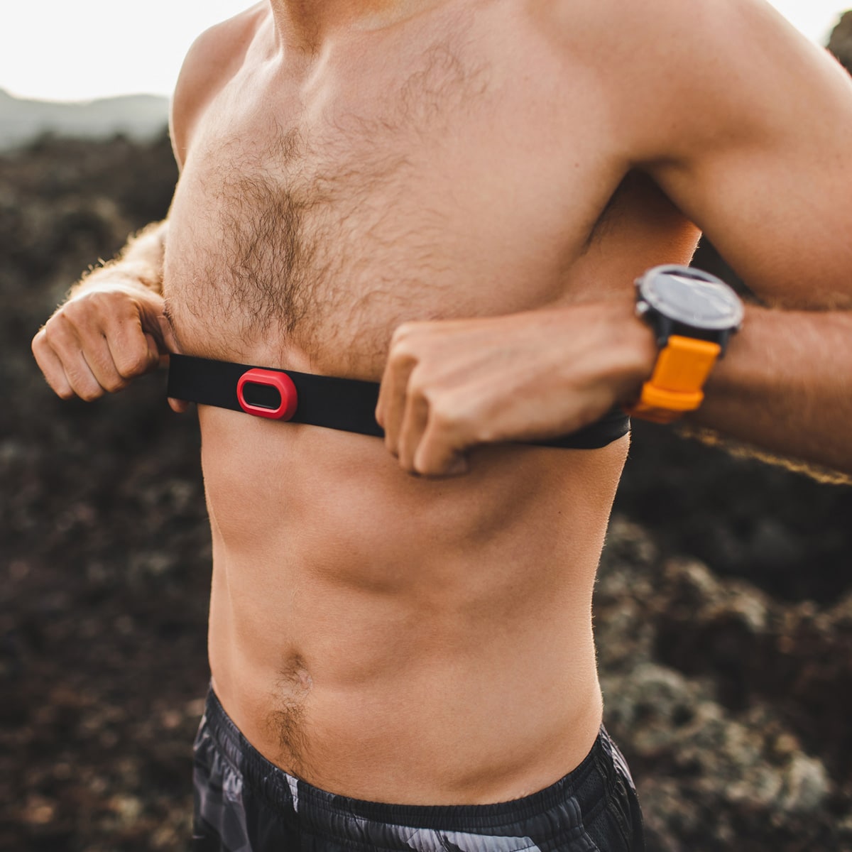 A shirtless man outdoors adjusts a heart rate monitor strap around his chest. Wearing a wristwatch, he stands against a rocky background.