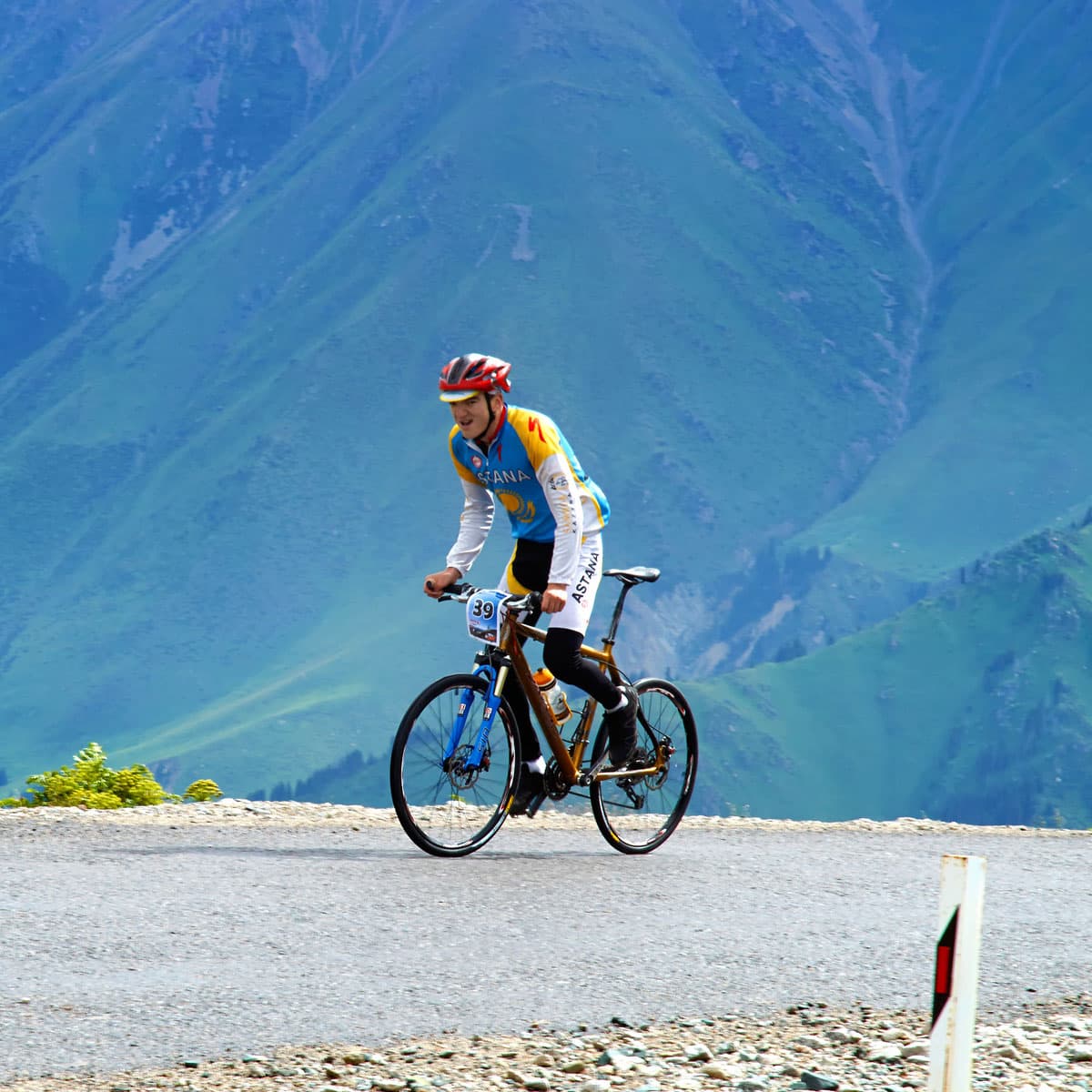 A cyclist wearing a helmet and race gear rides up a mountain road with green hills in the background.