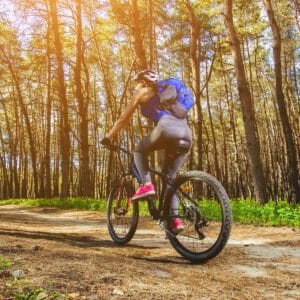 A cyclist wearing a helmet and backpack rides a mountain bike on a sunlit forest trail, with tall trees and a bright sky.