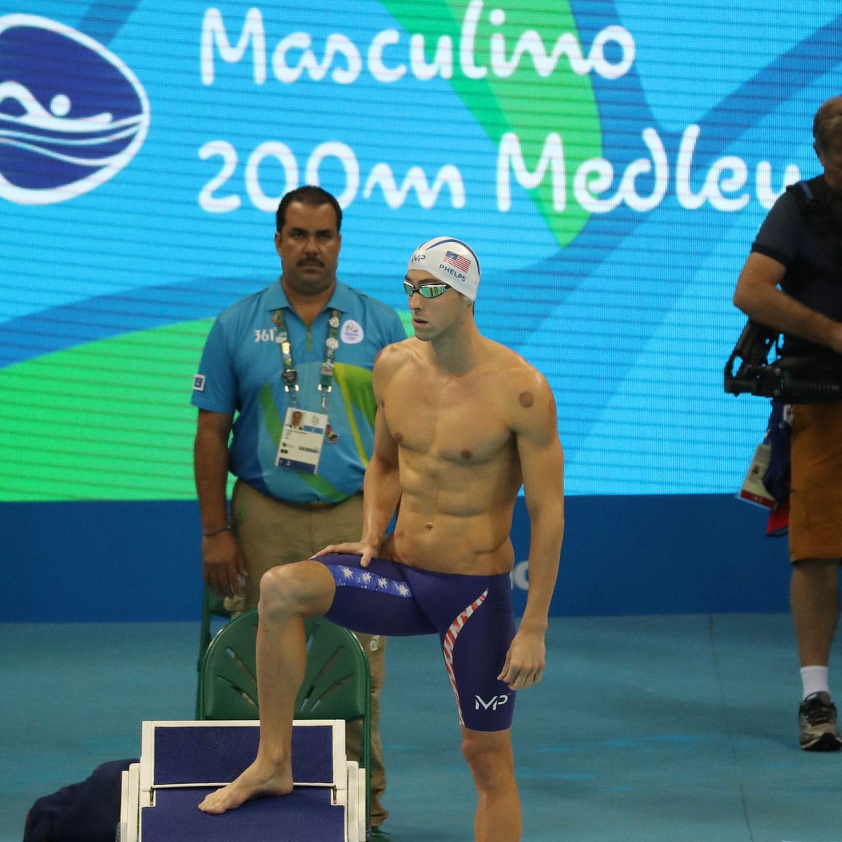 A male swimmer in goggles and a swim cap stands on a starting block, preparing for a race. An official is in the background, with a 200m Medley banner.
