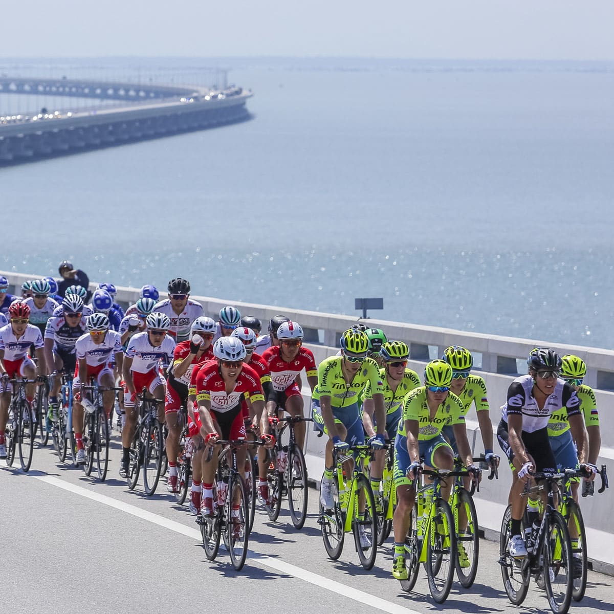 A large group of cyclists races along a coastal bridge with cross winds, with the sea in the background. This vibrant scene from captures the concept of a jam in grand tour cycling.