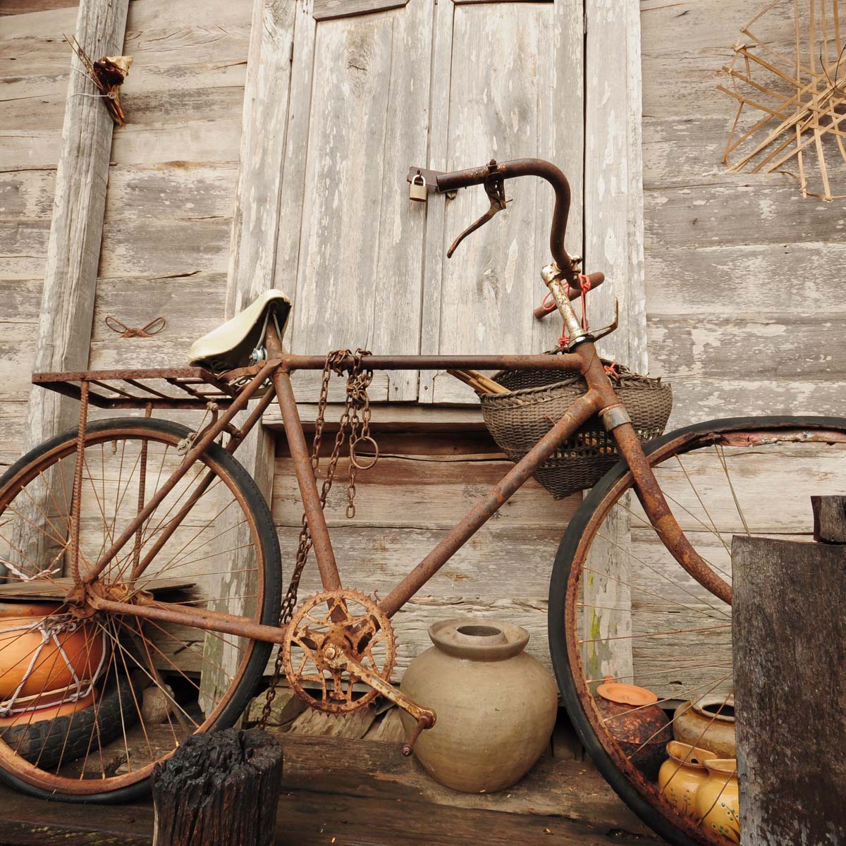 A rusty, broken bicycle leans against a weathered wooden wall. The handlebars and front wheel are twisted, and a basket hangs from the frame.