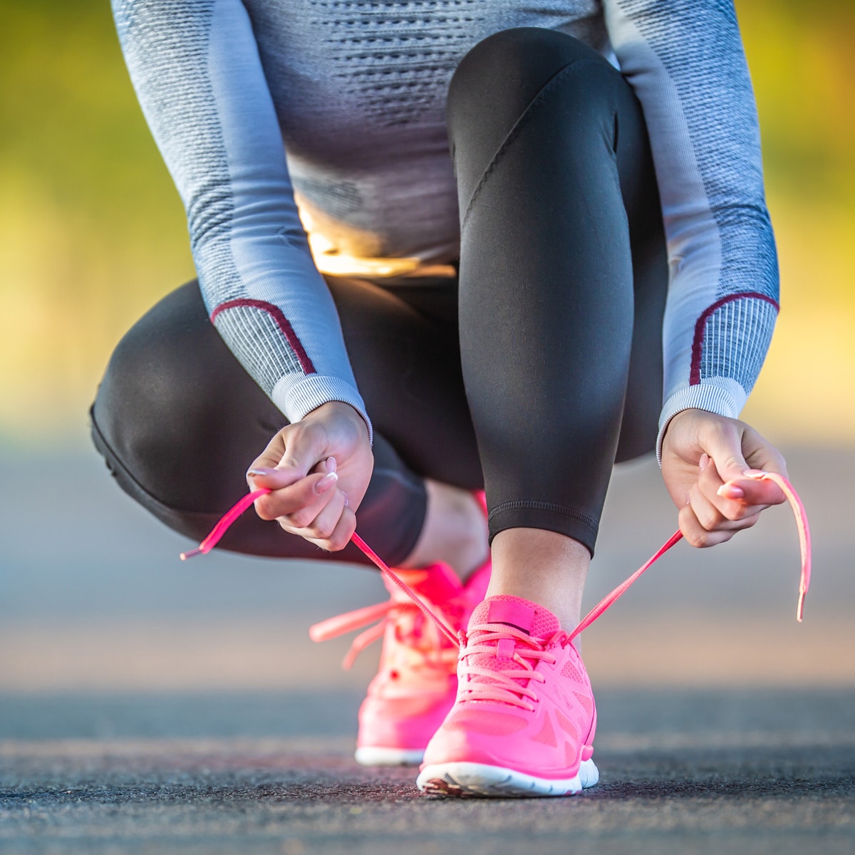 A runner wearing black leggings and a gray long-sleeve top is crouched down outdoors, tying the bright pink laces of her running shoes on a paved surface, resembling the concept of beginning a junk miles run.
