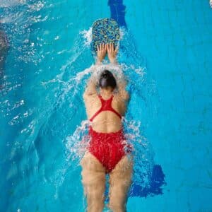 A swimmer in a red swimsuit uses a colorful kickboard in a pool, viewed from above. The swim cap and goggles are visible.