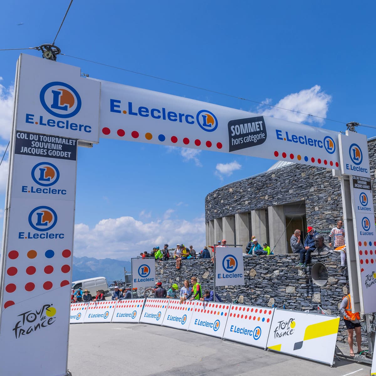 A large E.Leclerc-branded arch at a mountain stage finish line, with people gathered below. Stone building and mountains in the background under a blue sky.