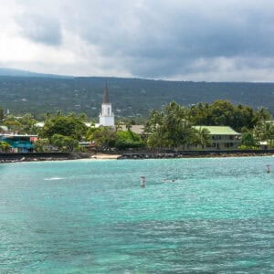 A coastal scene in Kailua-Kona, Hawaii, with turquoise water, palm trees, and a white church steeple set against lush green hills and a cloudy sky, blending the concept of the Ironman World Championship location with paradise.