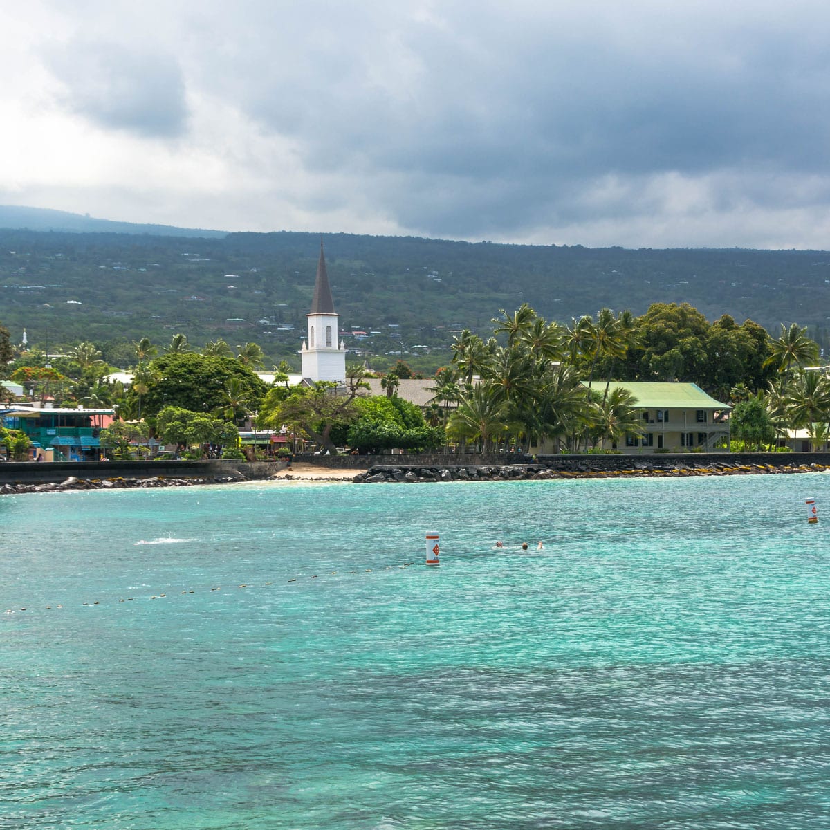 A coastal scene in Kailua-Kona, Hawaii, with turquoise water, palm trees, and a white church steeple set against lush green hills and a cloudy sky, blending the concept of the Ironman World Championship location with paradise.