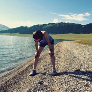 A female runner in athletic clothing leans forward, hands on knees, catching her breath on a gravel path beside a lake with mountains and blue sky.