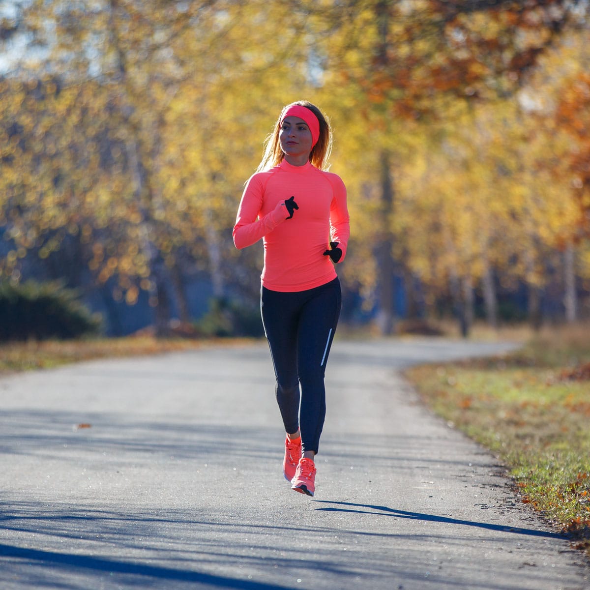 A woman in a bright pink long-sleeve top, headband, and black leggings enjoys a Long Slow Distance jog on a paved path through a park with autumn trees and fallen leaves in the background.