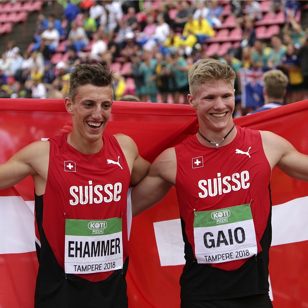 Two male Swiss athletes, smiling and celebrating with a Swiss flag after a race in a stadium with a national record broken. Wearing red Suisse jerseys and name bibs, Ehammer and Gaio showcase their triumph—moments of endurance and pride.