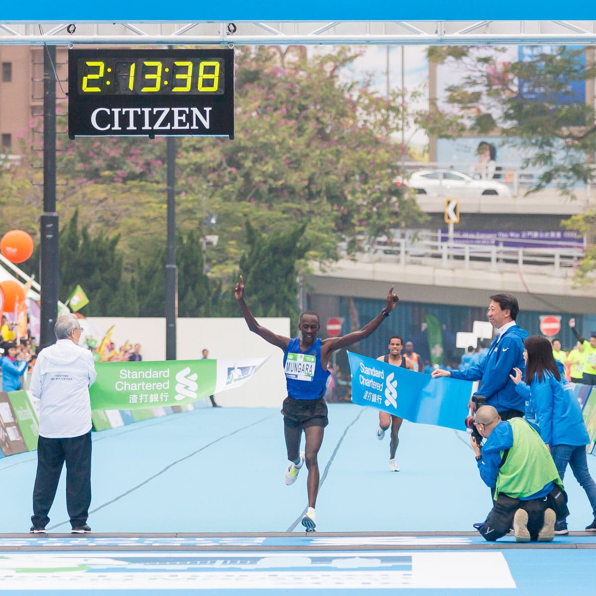 A marathon runner crosses the finish line with arms raised in victory. The digital clock above shows 2:13:38. Spectators and officials stand nearby.