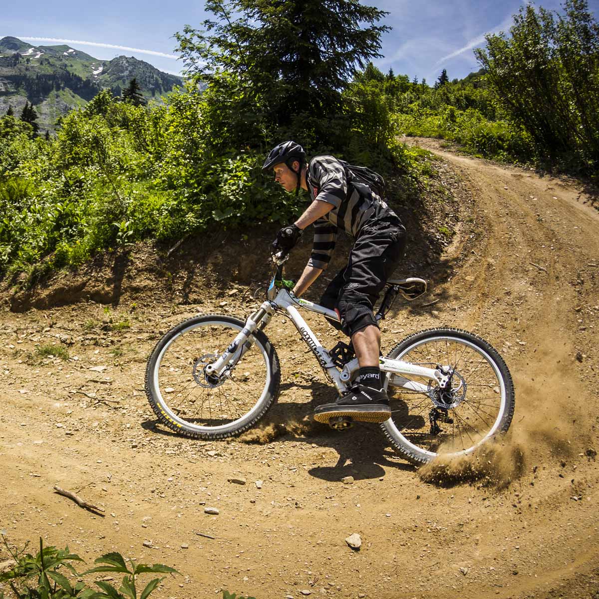 A mountain biker in full gear tackles a steep off-camber turn on a dirt trail, surrounded by trees and mountains under a clear blue sky.