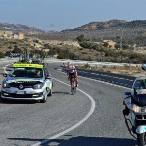 A cyclist rides uphill on a winding road, followed closely by a support car carrying spare bicycles. Rugged hills and dry terrain are visible in the background under a clear sky.