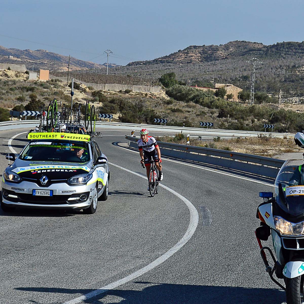 A cyclist rides uphill on a winding road, followed closely by a support car carrying spare bicycles. Rugged hills and dry terrain are visible in the background under a clear sky.