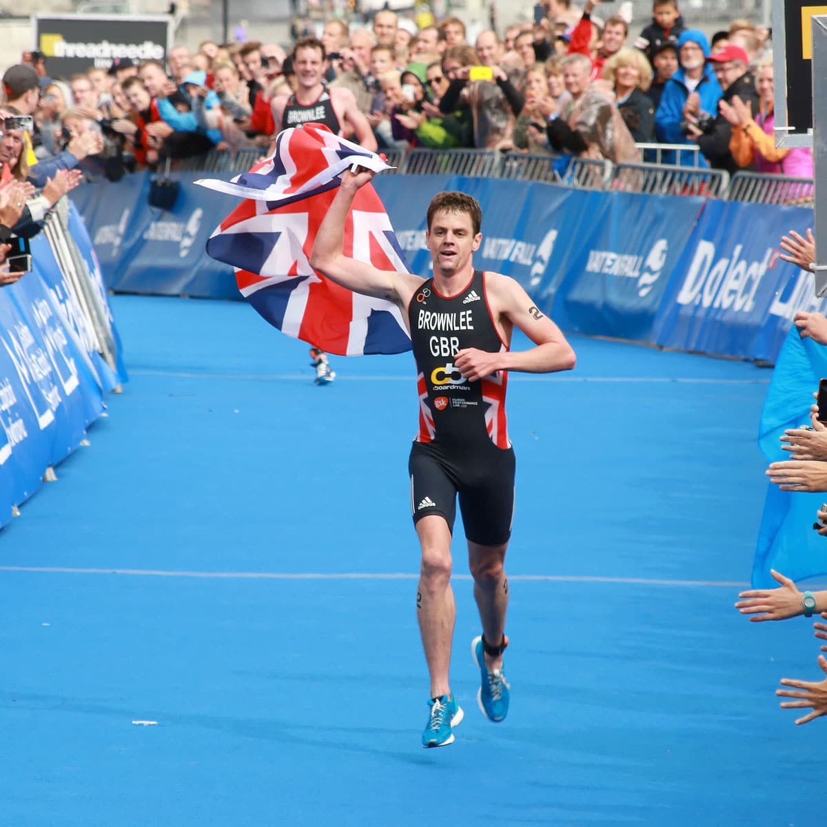 A triumphant male triathlete in a GBR suit runs toward the finish line, waving a British flag with cheering spectators looking on—an inspiring moment at this outdoor race event.