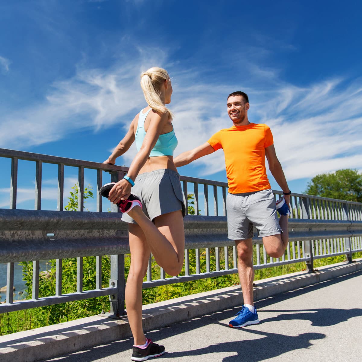 A man and woman in athletic wear stretch their quadriceps by holding their ankles while standing beside a metal railing outdoors on a sunny day, with blue sky and clouds in the background.