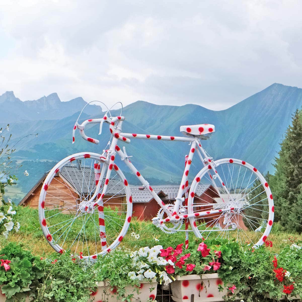A white bicycle with red polka dots sits on grass with flowers, in front of wooden cabins and tall mountains beneath a cloudy sky.