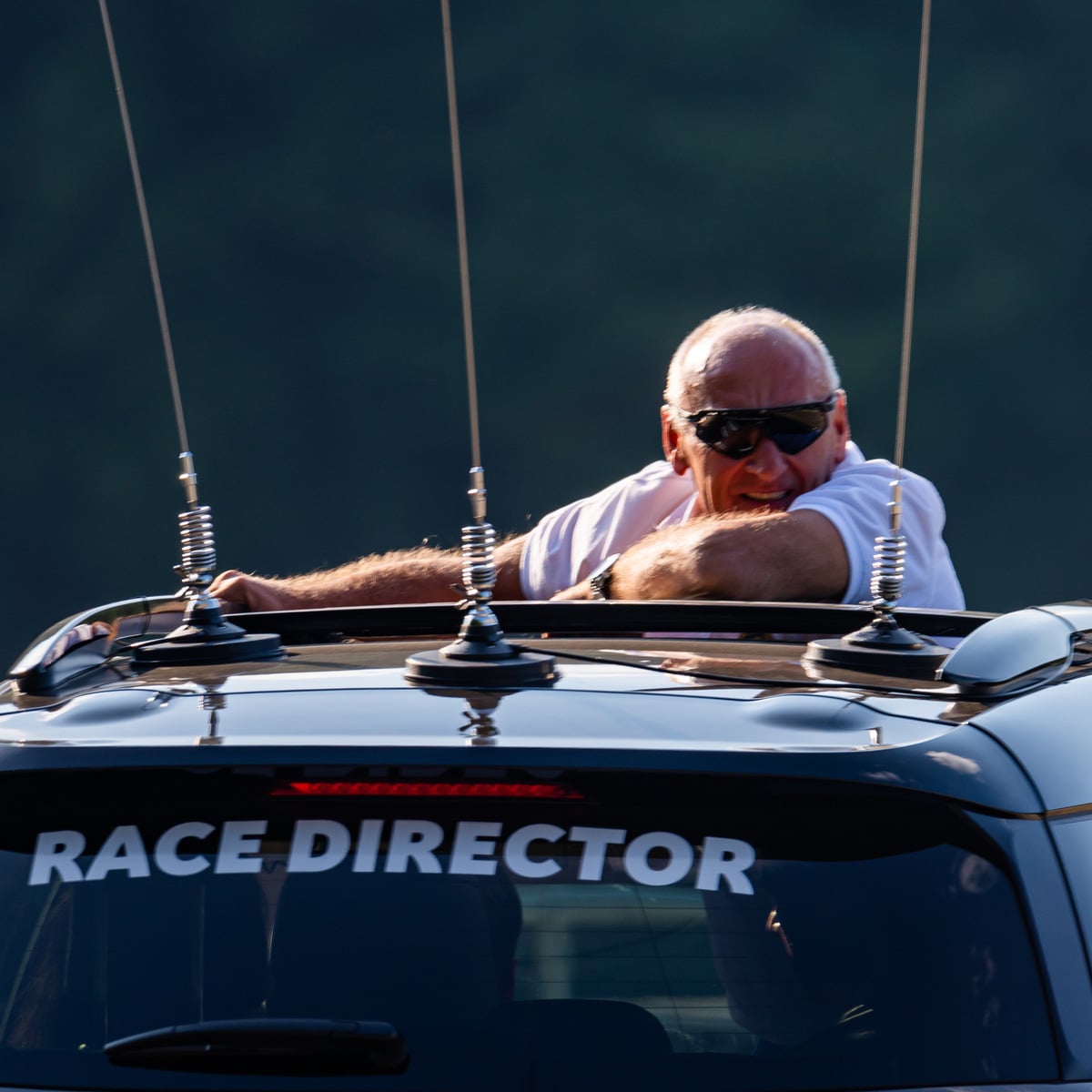 A Race Director wearing sunglasses leans out of the sunroof of a vehicle, gripping the roof with antennas in view. The background is blurred and out of focus.