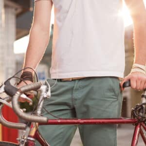 A person in a white shirt and green shorts stands with a red bicycle, holding the handlebars. Sunlight shines in the background.