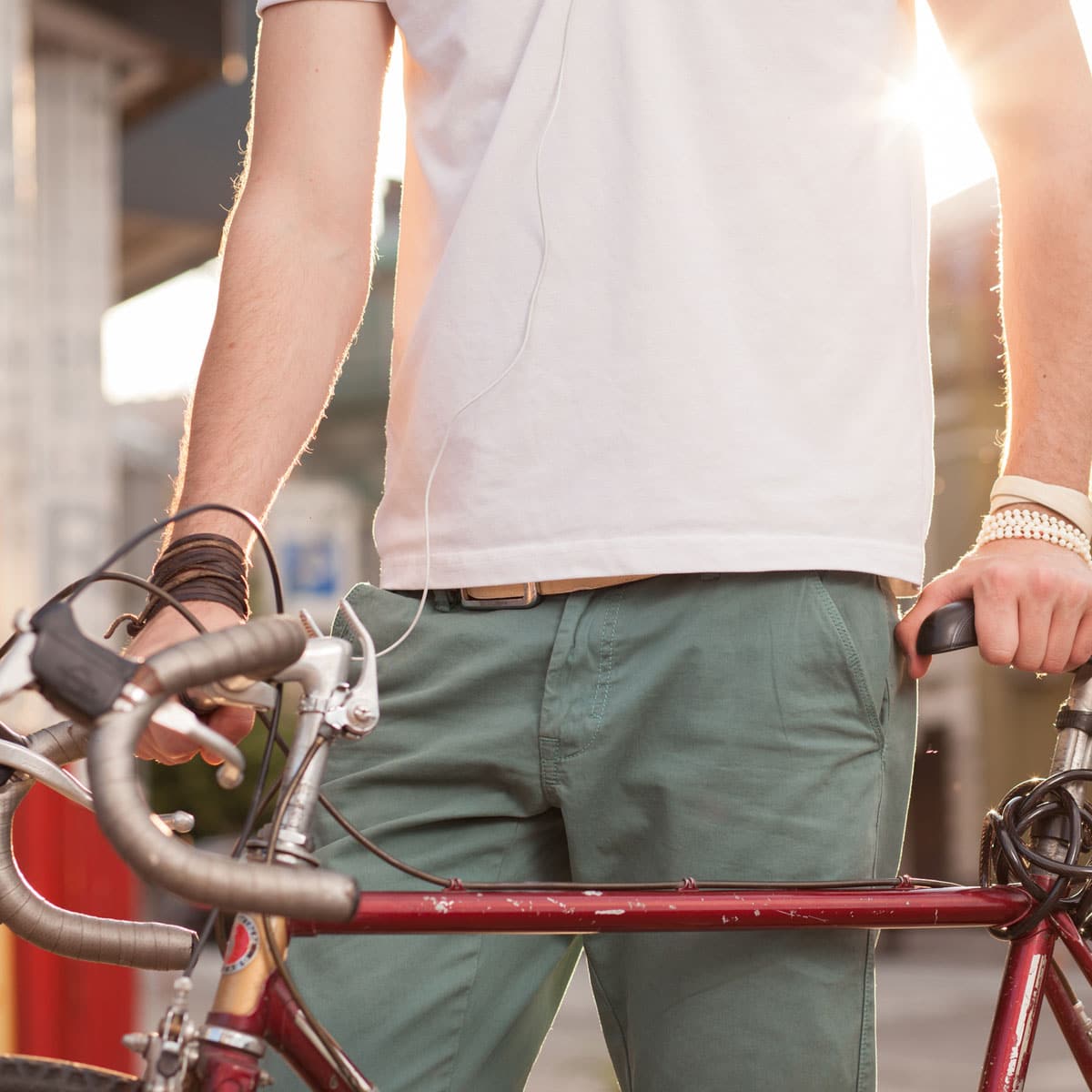 A person in a white shirt and green shorts stands with a red bicycle, holding the handlebars. Sunlight shines in the background.