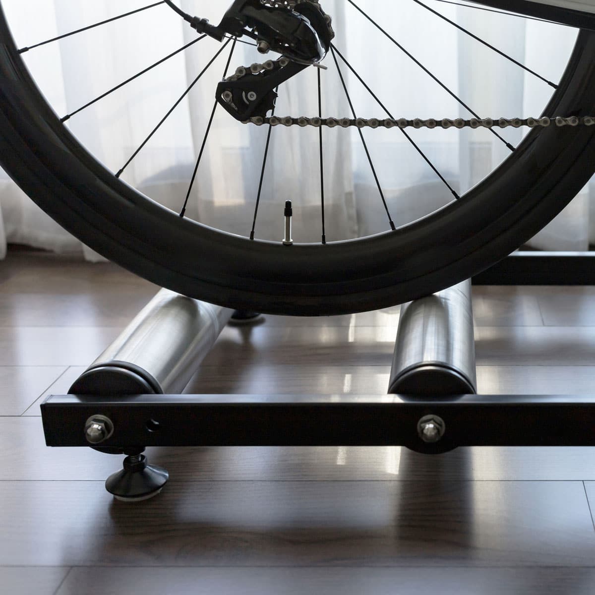 A close-up of a bicycle rear wheel on indoor rollers, positioned in front of a window with sheer curtains.