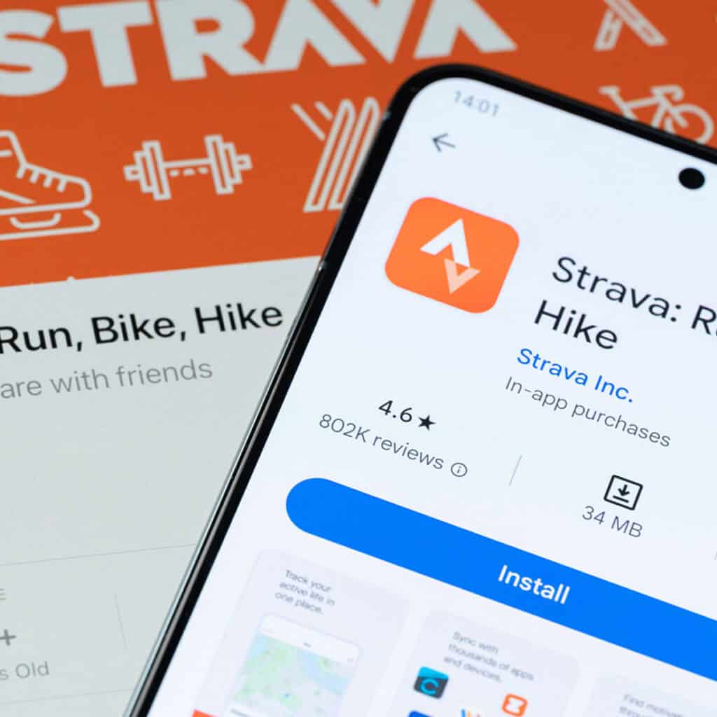A smartphone displays the Strava app screen, showing its rating and reviews. In the background, a Strava promotional graphic with fitness icons and the Strava logo is partially visible.