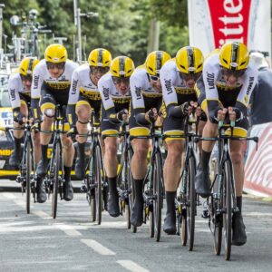 A professional cycling team in matching yellow and black helmets and gear rides closely together in a team time trial on a tree-lined road, with support cars following behind.
